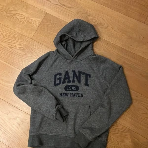 Grå hoodie från GANT - Säljer en snygg och bekväm grå hoodie från GANT med tryck '1949 New Haven' på bröstet. Perfekt för en avslappnad stil och passar bra till både jeans och joggers. Den har en klassisk passform med långa ärmar och en mysig huva. Perfekt för höst och vinter!