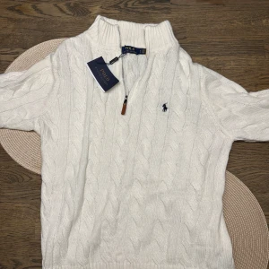 Polo tröja  - Säljer en snygg vit stickad tröja från Ralph Lauren med klassiskt kabelstickat mönster. Tröjan har en dragkedja framtill och den ikoniska loggan broderad på bröstet. Perfekt för kyliga dagar och ger en stilren look. Passar både till vardags och lite finare tillfällen. Tröjor är köpt på NK och är i i pricip nytt skick. Nypris: 2600kr