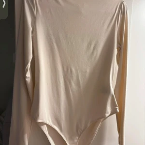 Beige långärmad body från Zara - Säljer en stilren beige body från Zara i storlek M. Den är långärmad och har en mjuk, stretchig passform som är perfekt för både vardag och fest. Materialet känns skönt mot huden och den neutrala färgen gör den lätt att matcha med olika outfits.