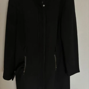 Svart kappa med dragkedjor - Snygg svart kappa i mycket bra skick. Den har långa ärmar och stilrena dragkedjor vid fickorna. Perfekt för höst och vinter, och funkar både till vardags och fest. En klassisk och tidlös stil som passar till det mesta.