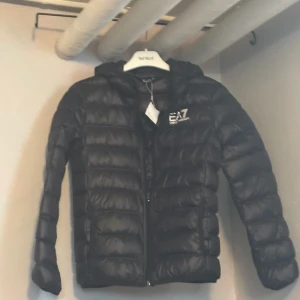 Svart dunjacka från EA7 - Säljer en snygg svart dunjacka från EA7. Den är perfekt för kalla dagar med sin pufferstil och har en praktisk huva. Jackan har en dragkedja framtill och är i mycket bra skick. Perfekt för både höst och vinter!