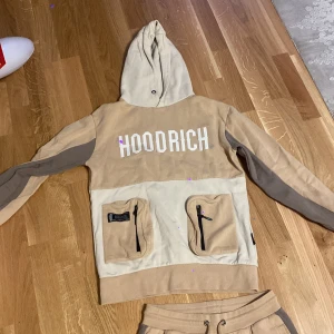 Beige och brun hoodie från Hoodrich - Säljer en snygg beige och brun hoodie från Hoodrich. Den har en stor logga på bröstet och praktiska fickor framtill. Perfekt för en avslappnad stil och passar bra till höst och vår. Hoodien är i mycket bra skick och redo för nya äventyr!
