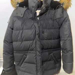 Svart dunjacka med pälsdetaljer - Säljer en varm och snygg svart boomerang dunjacka med pälsdetaljer på huvan. Jackan har långa ärmar och stängs med både dragkedja och knappar. Perfekt för kalla vinterdagar! 🖤strl M