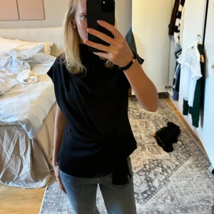 Svart topp med knytning - Säljer en stilren svart topp med knytning i midjan. Den är ärmlös och har en elegant, draperad design som passar perfekt för både vardag och fest. Materialet är satin och känns mjukt och bekvämt mot huden. Perfekt för att styla med jeans eller kjol för en chic look!