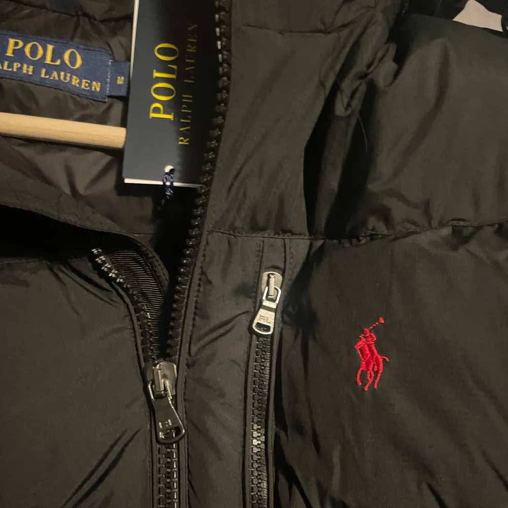 Säljer en svart dunjacka från Polo Ralph Lauren i storlek M. Jackan har en klassisk pufferstil med dragkedja och en liten röd logga på bröstet. Perfekt för kalla vinterdagar med sin varma och bekväma passform. Huvan ger extra skydd mot väder och vind. . Jackor.