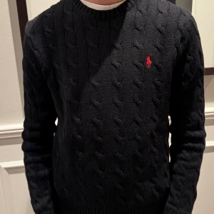 Svart stickad tröja från Polo Ralph Lauren - Säljer en snygg svart stickad tröja från Polo Ralph Lauren. Den har ett klassiskt flätmönster och en liten röd logga på bröstet. Den är i riktigt fint skick, modellen på bilden är 168cm och 52 kg