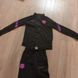 Säljer ett svart träningsset från Nike med Barcelona-logga. Setet har en jacka med dragkedja och matchande byxor. Detaljer i lila och rosa på ärmar och ben ger en cool look. Perfekt för fotbollsträning eller vardagsbruk. Dri-FIT material för bästa komfort.Finns ett lite hål i byxan med det syns knappt. Storlek L barn 147-157