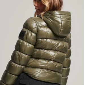 Olivgrön pufferjacka med huva - Säljer en snygg olivgrön pufferjacka med huva. Jackan har en glansig finish och är perfekt för kyliga dagar. Den har en dragkedja framtill och är i mycket bra skick. Perfekt för höst och vinter!