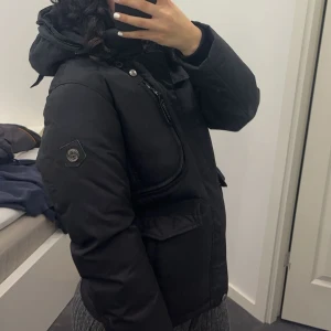 Svart vinterjacka med pälsdetalj - Säljer en svart vinterjacka i mycket bra skick. Den har en avtagbar luva och äkta beige päls, pälsen är köpt ifrån själva hollies butik och praktiska fickor med dragkedja. Perfekt för kalla vinterdagar! Jackan är varm och bekväm, idealisk för både vardag och utomhusaktiviteter.