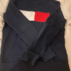  Tommy Hilfiger tröja  - Säljer en snygg mörkblå tröja från Tommy Hilfiger med deras ikoniska logga i rött och vitt på bröstet. Tröjan är i bra skick och perfekt för både höst och vinter. Den har långa ärmar och en klassisk passform. Perfekt för en stilren och avslappnad look!