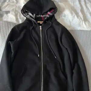 Säljer en snygg svart hoodie från Burberry i bra skick. Den har en dragkedja framtill och en klassisk rutig mönster på insidan av huvan. Perfekt för höst och vår, med långa ärmar och en bekväm passform. Passar både till vardags och lite finare tillfällen.