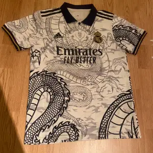 Säljer en unik Real Madrid fotbollströja från Adidas med ett coolt drakmönster. Tröjan är kortärmad och har en stilren krage. Den är i mycket bra skick och har spelarnamnet 'Vinicius Jr' och nummer 20 på ryggen. Perfekt för samlare eller fans av laget! Pris går att diskutera!