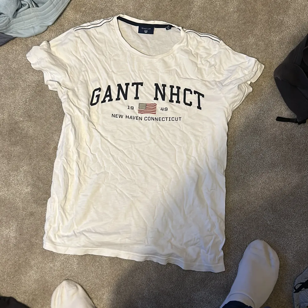 Säljer en vit t-shirt från Gant med tryck 'GANT NHCT' och en amerikansk flagga. Den har en liten defekt längst ner. Perfekt för en avslappnad stil eller sommarhäng. Passar bra till jeans eller shorts!. T-shirts.