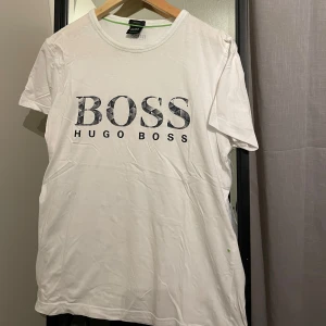 Vit t-shirt från Hugo Boss - Säljer en stilren vit t-shirt från Hugo Boss i bomull, väldigt bra skick samt storlek S. Den har en BOSS-logga på framsidan i en cool, mönstrad stil och en mindre logga på baksidan. Perfekt för en casual look eller att klä upp med en snygg jacka. Passar både till vardags och fest!