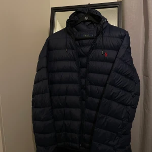 Dunjacka från Polo Ralph Lauren - Säljer en snygg dunjacka från Polo Ralph Lauren i storlek M. Jackan är i nyskick och har en huva och dragkedja framtill, samt två praktiska fickor med dragkedjor. Den är quiltad och perfekt för kyliga dagar. Den röda broderade loggan på bröstet ger en stilren touch. Perfekt för höst och vinter, håller dig varm och stilren. Passar både till vardags och finare tillfällen, nypris 5000kr 🖤