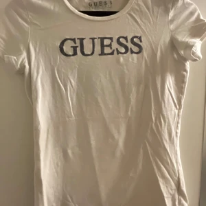 Vit t-shirt från Guess - Säljer en snygg vit t-shirt från Guess i bra skick. Den har korta ärmar och ett stort Guess-tryck på framsidan. På baksidan finns ett coolt frågetecken-tryck. Perfekt för en casual look eller att styla upp med accessoarer. Passar till alla tillfällen!