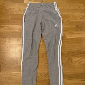 Grå mjukisbyxor från Adidas - Säljer ett par grå mjukisbyxor från Adidas i storlek S. Byxorna har de klassiska tre vita ränderna längs sidorna och en elastisk midja med dragsko för bästa passform. Perfekta för en avslappnad dag eller träning. 🩶