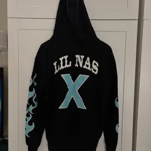 Cool svart hoodie från H&M Divided med blåa flammor längs ärmarna och texten 'LIL NAS X' på ryggen. Framsidan har texten 'PANINI' i gult. 