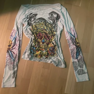 Vit långärmad tröja med dödskallemotiv - Cool vit långärmad tröja med ett stort, färgglatt dödskallemotiv och spindlar. Tröjan har en v-ringad hals och är perfekt för en unik och edgy stil. Den har också detaljer vilket ger en extra touch av attityd.
