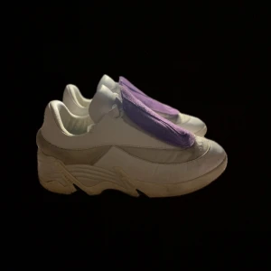 Raf Simon skor - Säljer ett par vita sneakers med lila detaljer. Skorna har en chunky sula och är i bra skick, men med en liten fläck/skavank/hål framtill. Perfekta för en avslappnad stil och passar till både vardag och fest.