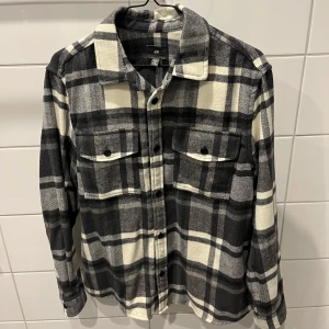 Svartvit rutig overshirt från H&M - Säljer en snygg och stilren svartvit rutig overshirt från H&M. Perfekt för lager-på-lager under höst och vår. Skjortan har långa ärmar och knappar framtill samt två bröstfickor med knappar. Den är i mycket bra skick och passar både till vardags och lite mer uppklädda tillfällen.