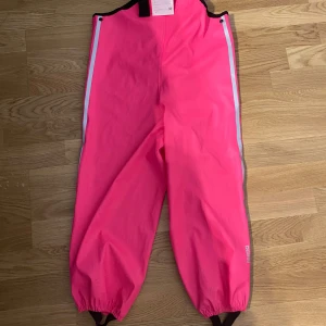 Rosa regnbyxor st. 116 - Säljer ett par knallrosa regnbyxor  i bra skick. De har elastiska muddar vid bensluten och reflexdetaljer längs sidorna för extra synlighet. Perfekta för regniga dagar och håller dig torr och bekväm. Passar bra för utomhusaktiviteter!