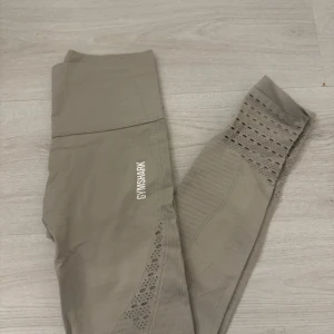 Grå leggings från Gymshark - Säljer ett par snygga grå leggings från Gymshark i storlek XS. De är högmidjade och har en tight passform, perfekt för träning. Materialet är stretchigt och bekvämt med snygga hålmönster längs benen för extra stil. Perfekta för gymmet eller yoga!