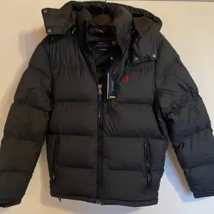 Säljer en helt oanvänd dunjacka från Polo Ralph Lauren i storlek S, men passar även för dig som brukar ha M. Jackan har en avtagbar huva och dragkedja framtill. Den är perfekt för kalla vinterdagar och har en klassisk design med det ikoniska röda logotypbroderiet på bröstet. Håll dig varm och stilren i vinter! Nypriset ligger på 5800kr och tagen finns kvar!! Skicka DM om du vill ha fler bilder eller se kvittot!! Tveka inte vid funderingar.