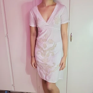 Vit kortklänning med broderi - Säljer en söt vit kortklänning med v-ringning och korta ärmar. Klänningen har vackra broderade detaljer i beige som ger en romantisk känsla. Perfekt för sommarens fester eller en avslappnad dag på stan. Materialet känns lätt och luftigt.