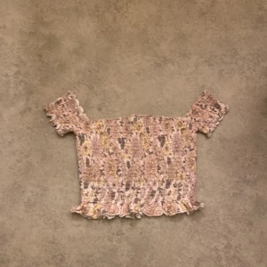 Mönstrad offshoulder topp från BikBok - Säljer en superfin offshoulder topp från BikBok i storlek M. Den har ett unikt mönster i rosa och beige toner med volangdetaljer vid ärmslut och nederkant. Perfekt för sommardagar eller en festlig kväll!