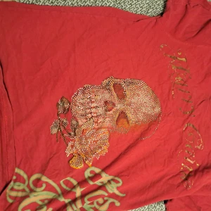 Röd t-shirt med dödskalle och örn - Cool röd t-shirt med ett stort tryck av en dödskalle och en örn. Dödskallen är dekorerad med glittriga detaljer och det finns även en färgglad design med en blå och orange fjäril. Perfekt för en unik och edgy stil!