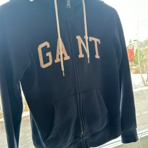 Mörkblå hoodie från GANT - Säljer en snygg mörkblå hoodie från GANT med dragkedja och stora fickor framtill. Den har en klassisk logga i vitt på bröstet och justerbara snören i huvan. Perfekt för kyliga dagar eller som ett stilrent lager under jackan. Passar både till vardags och avslappnade tillfällen.