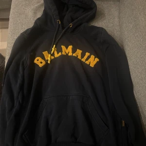 Mörk blå hoodie från Balmain - Säljer en snygg svart hoodie från Balmain med gul text på bröstet. Den har en klassisk känguruficka och justerbar huva med snören. Perfekt för en casual look eller att mysa i hemma. Passar bra till både jeans och andra . Använd några gånger.
