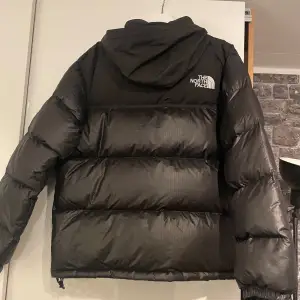 Säljer en svart dunjacka från The North Face i mycket bra skick. Jackan är perfekt för kalla dagar med sitt Eco Down-material som håller dig varm. Den har en dragkedja framtill och en huva för extra skydd. Loggan syns både fram och bak. Perfekt för vintern! Jackan är i strlk XL men passar även L. Kom med pris förslag!