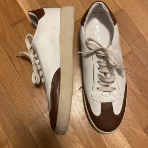 Vita och bruna skor från Brawn's - Säljer ett par snygga skor från Brawn's i vitt och brunt. De är i skinn och har en klassisk design med snörning. Perfekta för både vardag och fest! Tillverkade i Italien, vilket garanterar hög kvalitet. Sulan är i en krämig beige färg som ger en fin kontrast.
