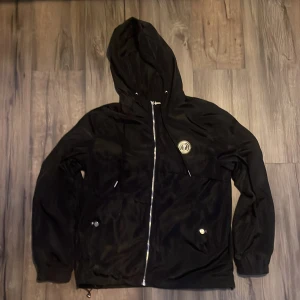 Svart jacka från Trapstar - Säljer en svart jacka från Trapstar i storlek M. Jackan har en dragkedja framtill och en huva. Den är dekorerad med ett märke på bröstet och texten 'Trapstar' på ryggen. Perfekt för höst och vår!