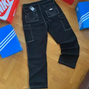 Helt Nya svarta cargobyxor från True Religion i storlek 32.  Nypris 1600kr, Mitt pris 899kr. Byxorna har en cool, distressed look med vita kontrastsömmar och flera praktiska fickor. Perfekta för en avslappnad och trendig stil. Passar bra till både vardag och fest! 