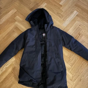 Svart parkajacka från Canada Goose - Säljer en svart parkajacka från Canada Goose i mycket bra skick. Jackan har en varm dunfyllning och en praktisk huva. Perfekt för kalla vinterdagar. Den har långa ärmar och en stilren design med dragkedja och knappar framtill. Pälskrage ingår                  Pris går att diskutera!