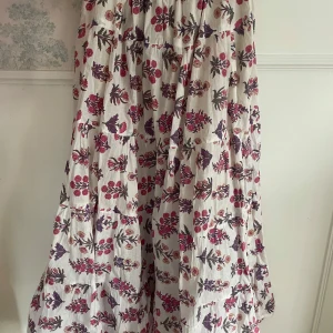 Blommig långkjol i bomull - Charmig långkjol med ett vackert blommönster i rött och lila på en vit bakgrund. Kjolen är tillverkad i bomull och har en luftig och bekväm passform, perfekt för varma dagar. Helt ny och köpt i Australien