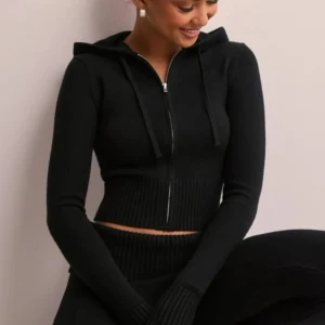 Svart croppad hoodie - Snygg svart croppad hoodie med dragkedja och huva. Tröjan tillhör det populära "lounge" settet från Nelly. Den har långa ärmar och ribbade detaljer vid midjan samt ärmsluten. Perfekt för en stilren och bekväm look.