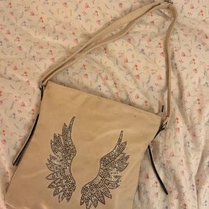 Beige axelväska med vingmotiv - Snygg beige axelväska med glittrigt vingmotiv på framsidan. Väskan har en dragkedja på baksidan och justerbar axelrem. Perfekt för att ge en unik touch till din outfit.