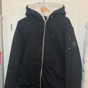 Svart jacka från Moose Knuckles - Säljer en svart jacka från Moose Knuckles med vit fuskpälsfoder i huvan. Jackan har dragkedja framtill och en snygg detalj på ärmen. Perfekt för kyliga dagar. Pris kan diskuteras vid snabb affär