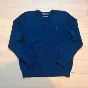 Blå ulltröja från Polo Ralph Lauren - Säljer en klassisk blå tröja från Polo Ralph Lauren i 100% lammull. Tröjan har en v-ringad design och långa ärmar, perfekt för kyligare dagar. Den ikoniska loggan är broderad på bröstet. Passar både till vardags och mer uppklädda tillfällen.