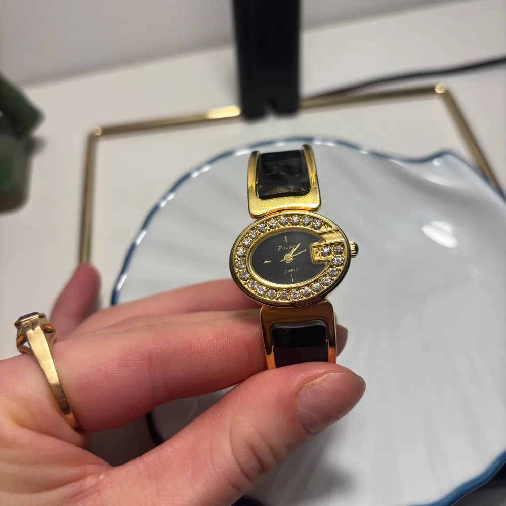 Snygg guldfärgad klocka med svart armband och glittrande stenar runt urtavlan. Perfekt accessoar för att lyfta din stil. Urtavlan är svart med guldfärgade detaljer. Fungerar!. Asusteet.