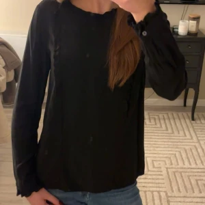 Svart långärmad blus - Snygg svart blus med långa ärmar och rund hals. Den har även små volang detaljer. Perfekt för en stilren look. Mycket bra skick