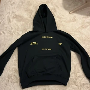 Kanye West (ye) hoodie - Säljer en svart hoodie med texten 'Jesus Is King' och 'Kanye West' i gult tryck. Hoodien har en klassisk design med känguruficka och huva. Perfekt för en avslappnad stil, i nytt skick. Stl L men passar S.