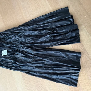 Svart långkjol - Snygg svart långkjol med glansigt material och veckad design. Perfekt för en elegant look. Skick 10/10 oanvänd tags kvar