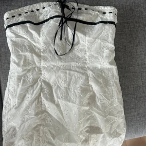 Vit axelbandslös klänning med spetsdetaljer - Charmig vit axelbandslös klänning med broderade blommor och spetsdetaljer. Svart band löper genom spetsen upptill och knyts framtill för en söt touch. Perfekt för somriga dagar.