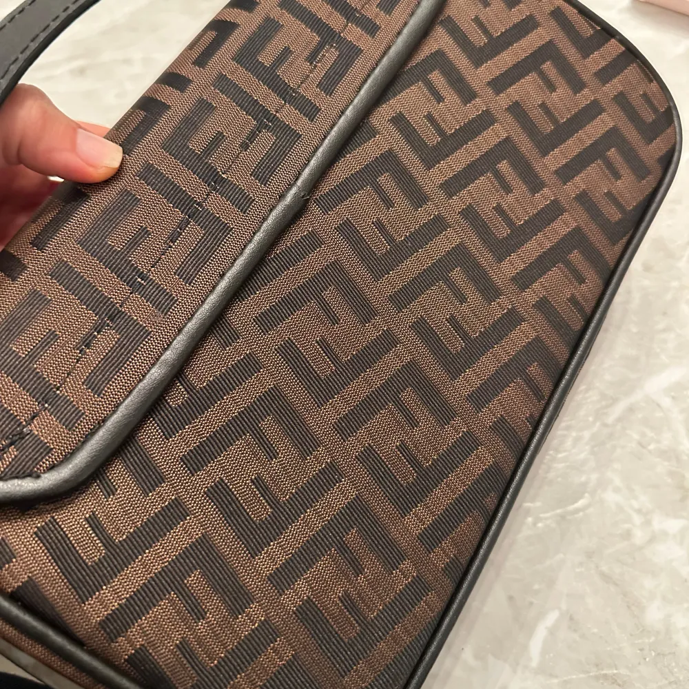 Snygg brun axelväska med monogrammönster och guldfärgad spänne. Väskan har en justerbar axelrem och är perfekt för att bära dina nödvändigheter med stil.. Laukut & Käsilaukut.