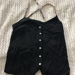 Svart linnetopp från Tommy Jeans - Snygg svart linnetopp från Tommy Jeans med knappar framtill och justerbara axelband med logotyp. Perfekt för en stilren och avslappnad look.